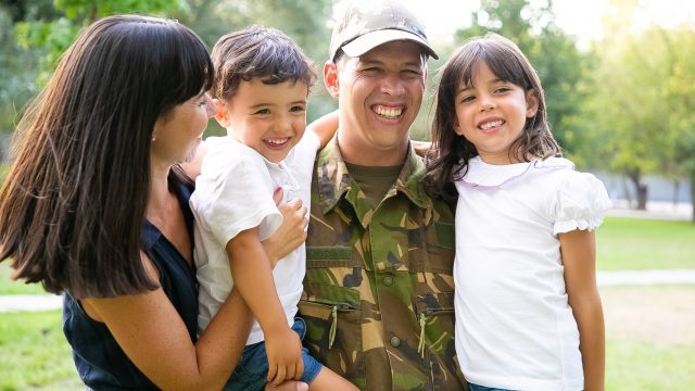 Happy military man posing for camera with his family, holding kids in arms, his wife hugging all of them and laughing. Medium shot. Family reunion or returning home concept